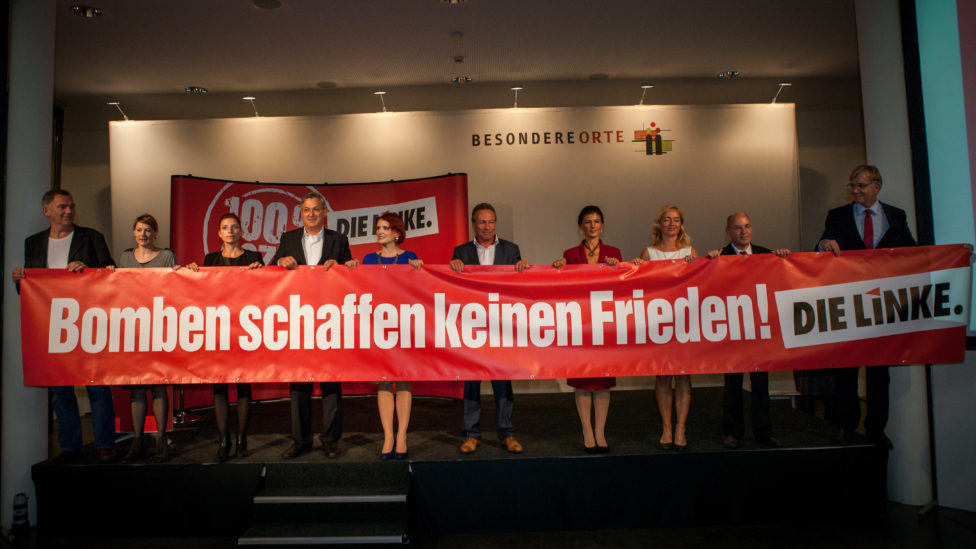 Jan van Aken, Nicole Gohlke, Diana Golze, Berbd Riexinger, Katja Kipping, Klaus Ernst, Dr. Sahra Wagenknecht, Caren Lay, Dr. Gregor Gysi und Dr. Dietmar Bartsch stehen auf einer Bühne und halten ein Transparent, auf dem "Bomben schaffen keinen Frieden! DIE LINKE."steht