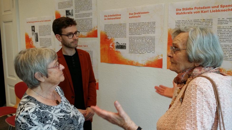 Tobias Bank und zwei ältere Damen stehen vor Ausstellungstafeln