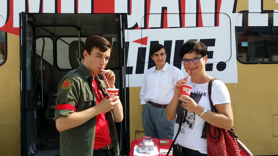 Aaron Birnbaum und Diana Bader stehen vor einem Doppeldecker-Bus und trinken mit Strohhalmen aus roten Pappbechern