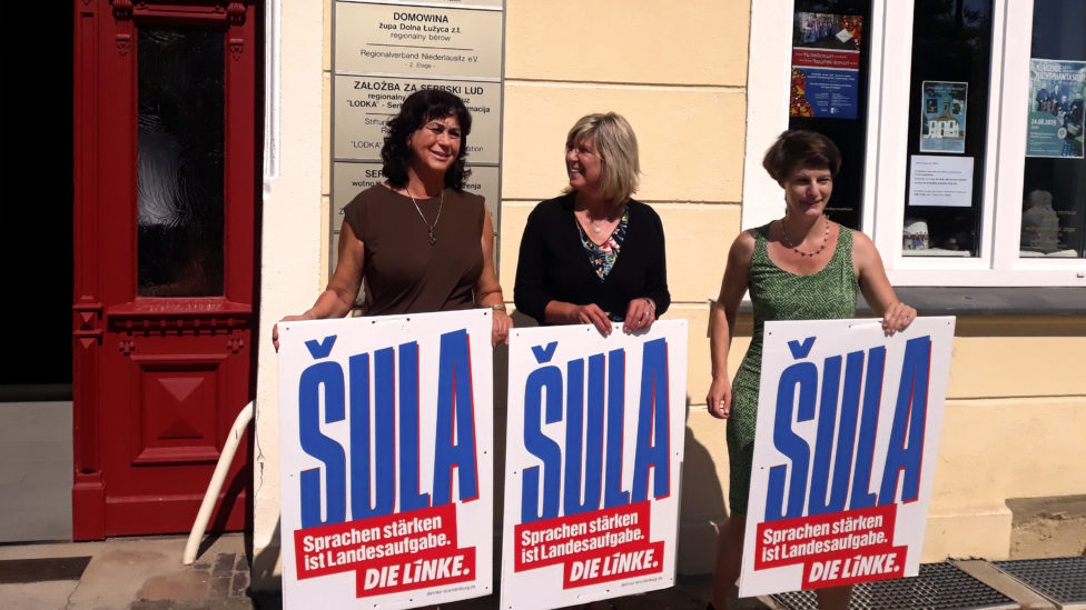Birgit Kaufhold, Kathrin Dannenberg und Monika von der Lippe stehen vor dem Wendischen Haus in Cottbus und halten Wahlplakate hoch, auf denen "Šula" steht