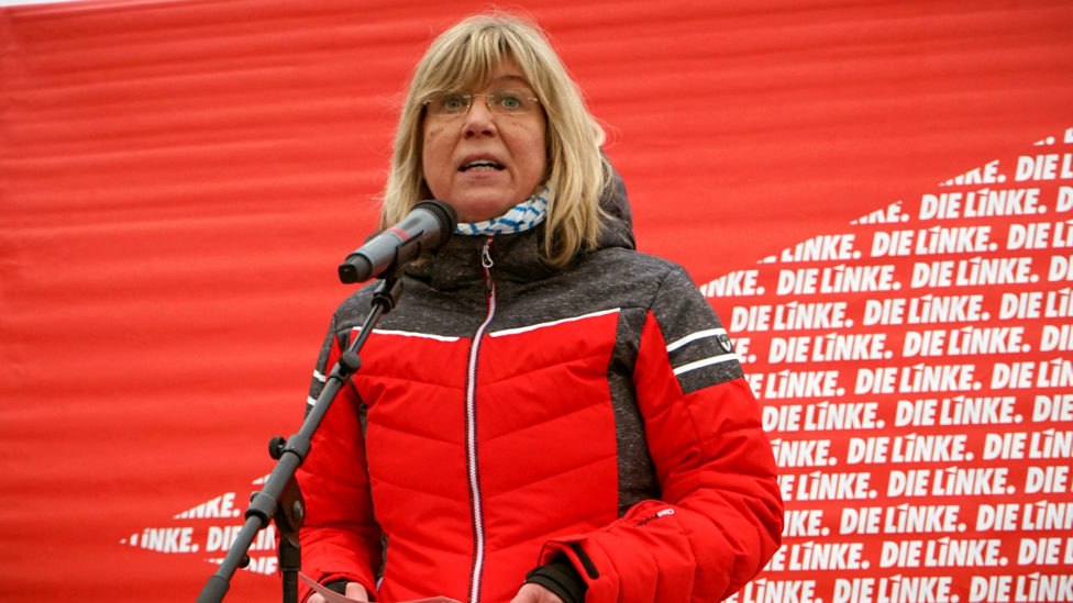 Kathrin Dannenberg steht am Mikrofon vor einem Transparent mit LINKE-Logos