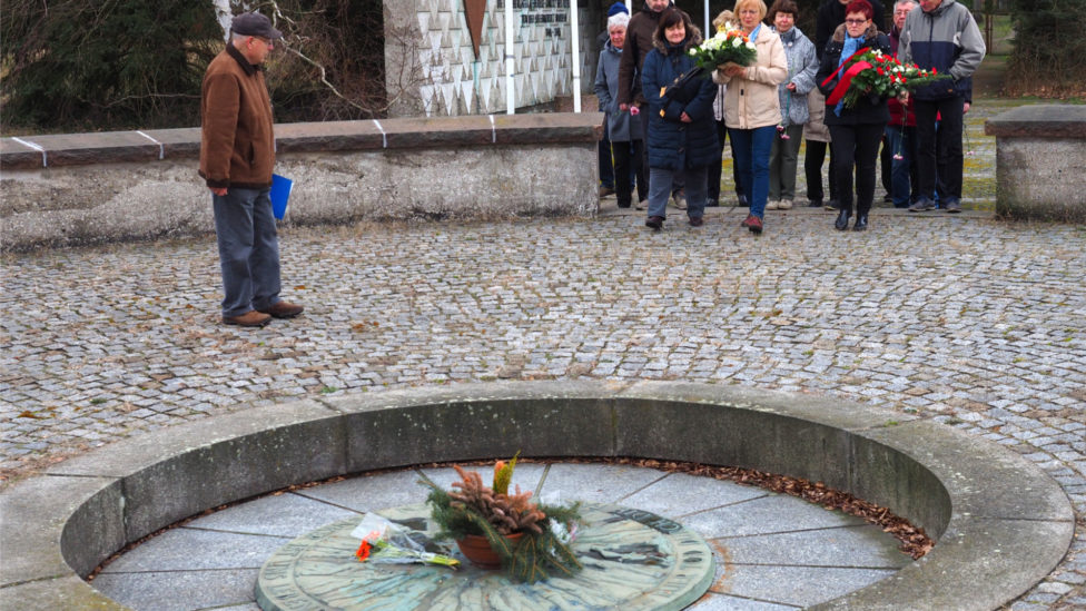 Eine Gruppe Menschen steht mit Gedenkkränzen vor dem Urnengrab in der Gedenkstätte für die Opfer des KZ Lieberose