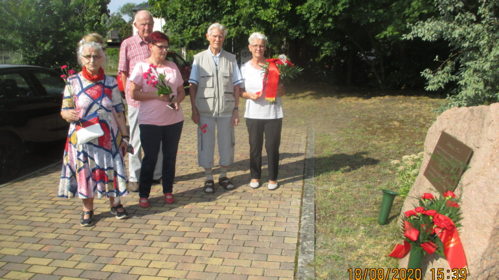 Eine Gruppe Menschen steht mit Blumen in den Händen vor einem Gedenkstein mit Gedenktafel