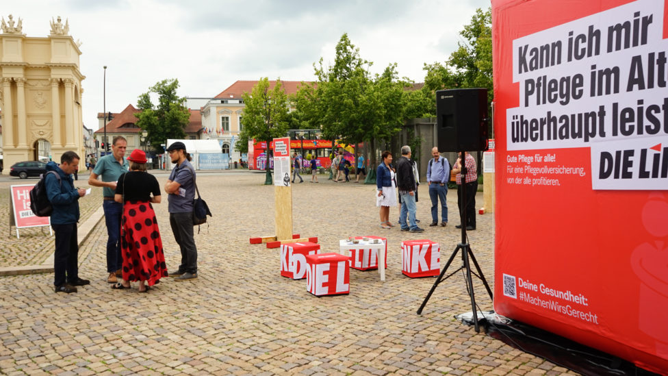 Luisenplatz in Potsdam, auf dem Platz steht ein 3x3 Meter großer aufblasbarer roter Würfel mit dem Logo der LINKEN und verschiedenen Aussagen zu den Themen Pflege und Gesundheit, um den Würfel herum stehen mehrere Menschen in Gruppen