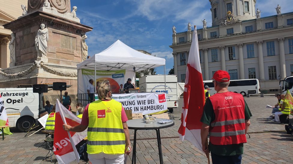 Menschen mit Warnwesten und Fahnen mit Logos der Gewerkschaften stehen vor dem Potsdamer Landtag, Norbert Müller steht unter einem Faltpavillon und spricht in ein Mikrofon