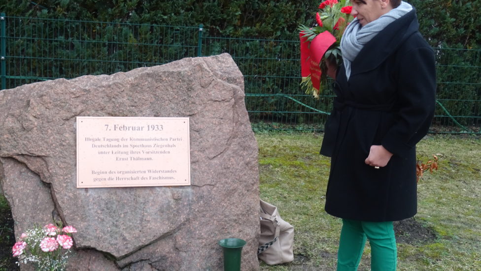 Katharina Slanina steht mit Blumen in den Händen vor dem Gedenkstein, an dem eine Gedenktafel befestigt ist