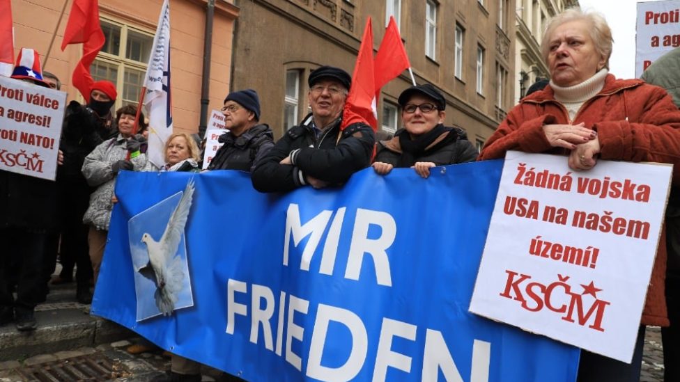 Teilnehmer der Kundgebung mit Schildern und roten Fahnen, mehrere Personen halten ein Transparent, auf das eine Friedenstaube sowie der Schriftzug "Mir - Frieden" gedruckt ist