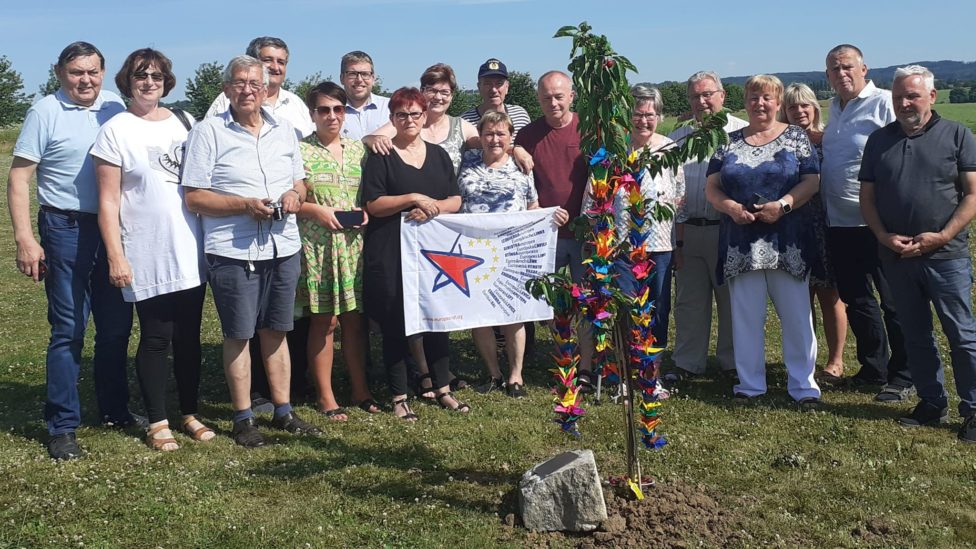 Gruppenfoto, bei dem Fotografierten hinter einem Gedenkstein stehen, neben dem ein Bäumchen mit Papiergirlanden gepflanzt wurde