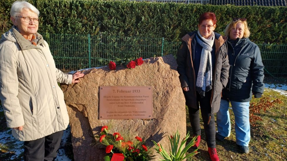 Mehrere Personen stehen am Gedenkstein, auf dem Blumen liegen