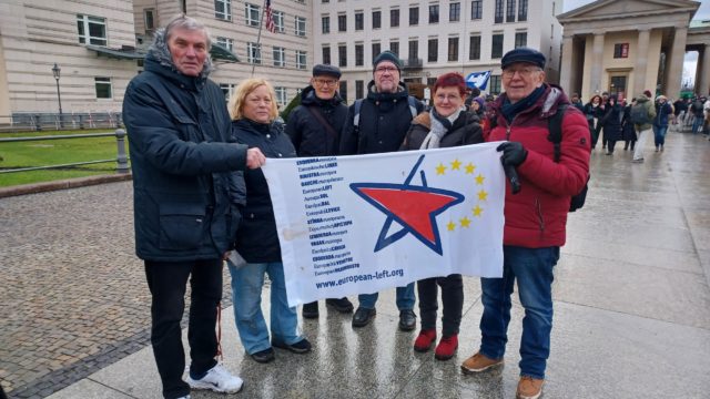 Mehrere Menschen stehen vor der US-Botschaft in Berlin und halten eine Fahne mit dem Logo der Europäischen Linken in den Händen